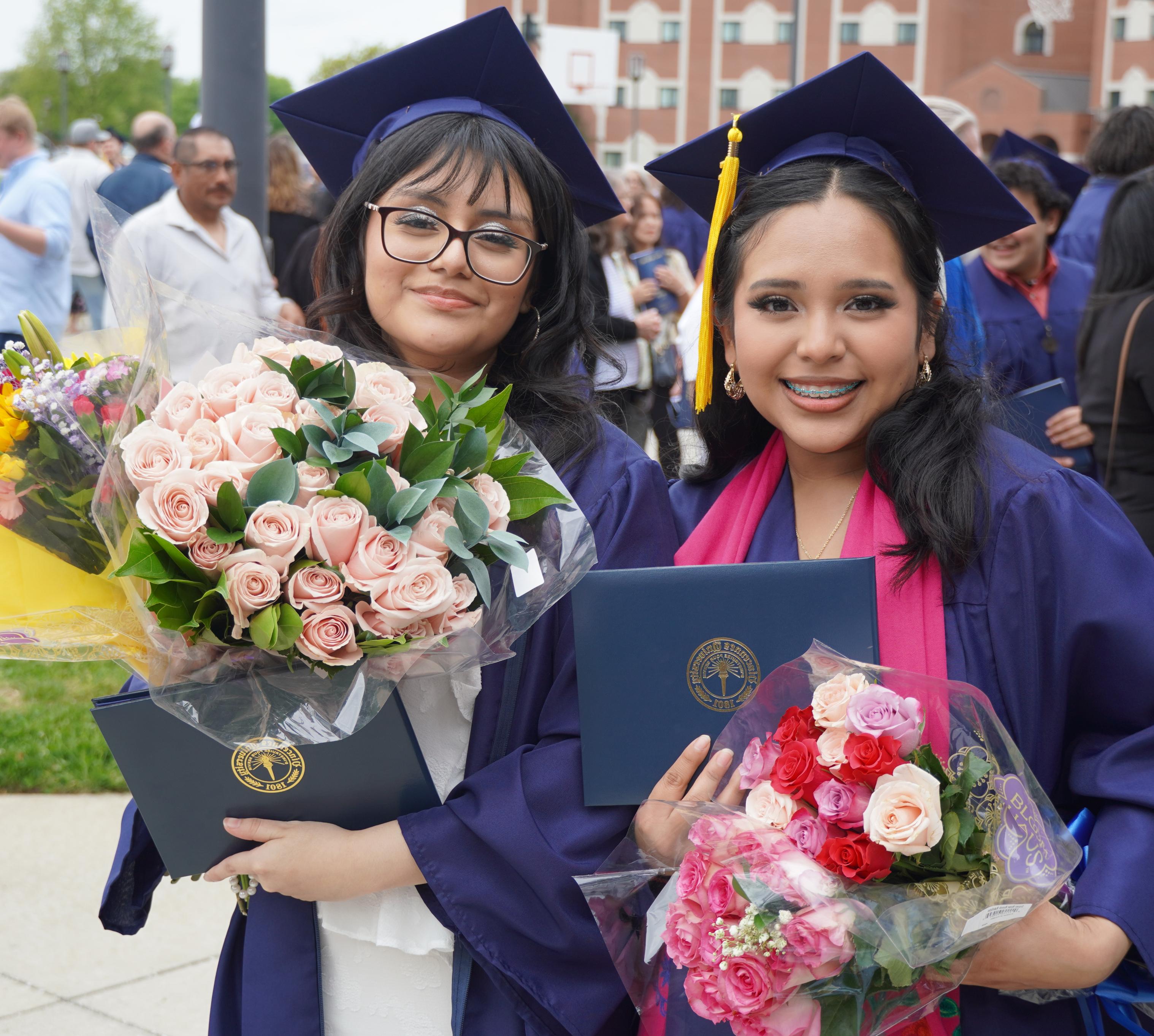 2 female wearing graduation caps and gowns while holding degree covers and flowers